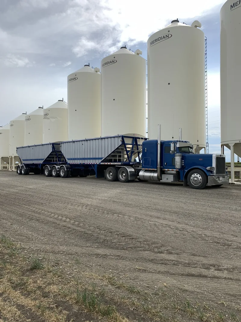 Blue grain truck and trailers beside a row of white storage bins